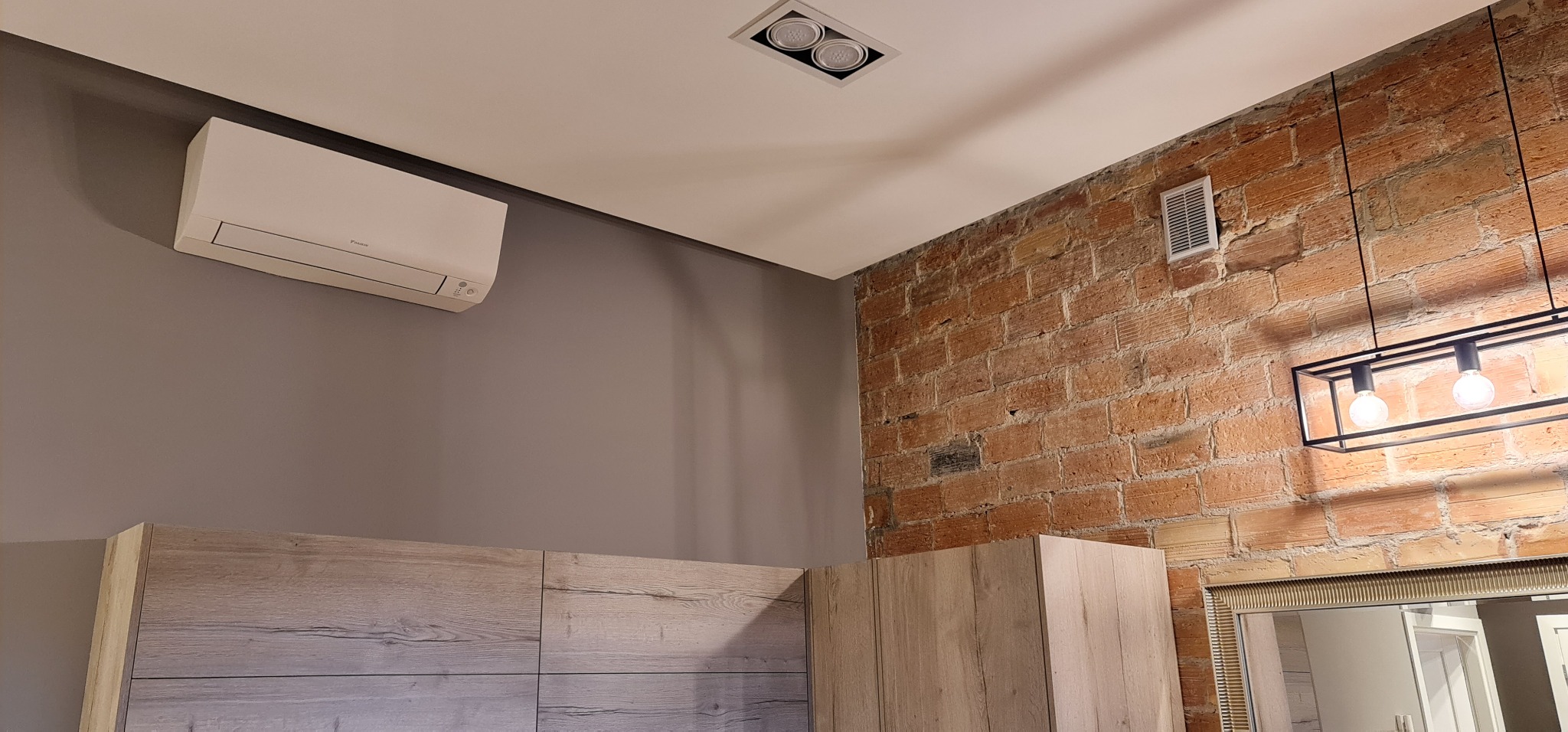Nowoczesny klimatyzator ścienny marki Daikin zamontowany na szarej ścianie w pomieszczeniu z ceglaną ścianą, oświetleniem sufitowym i meblami w kolorze jasnego drewna.