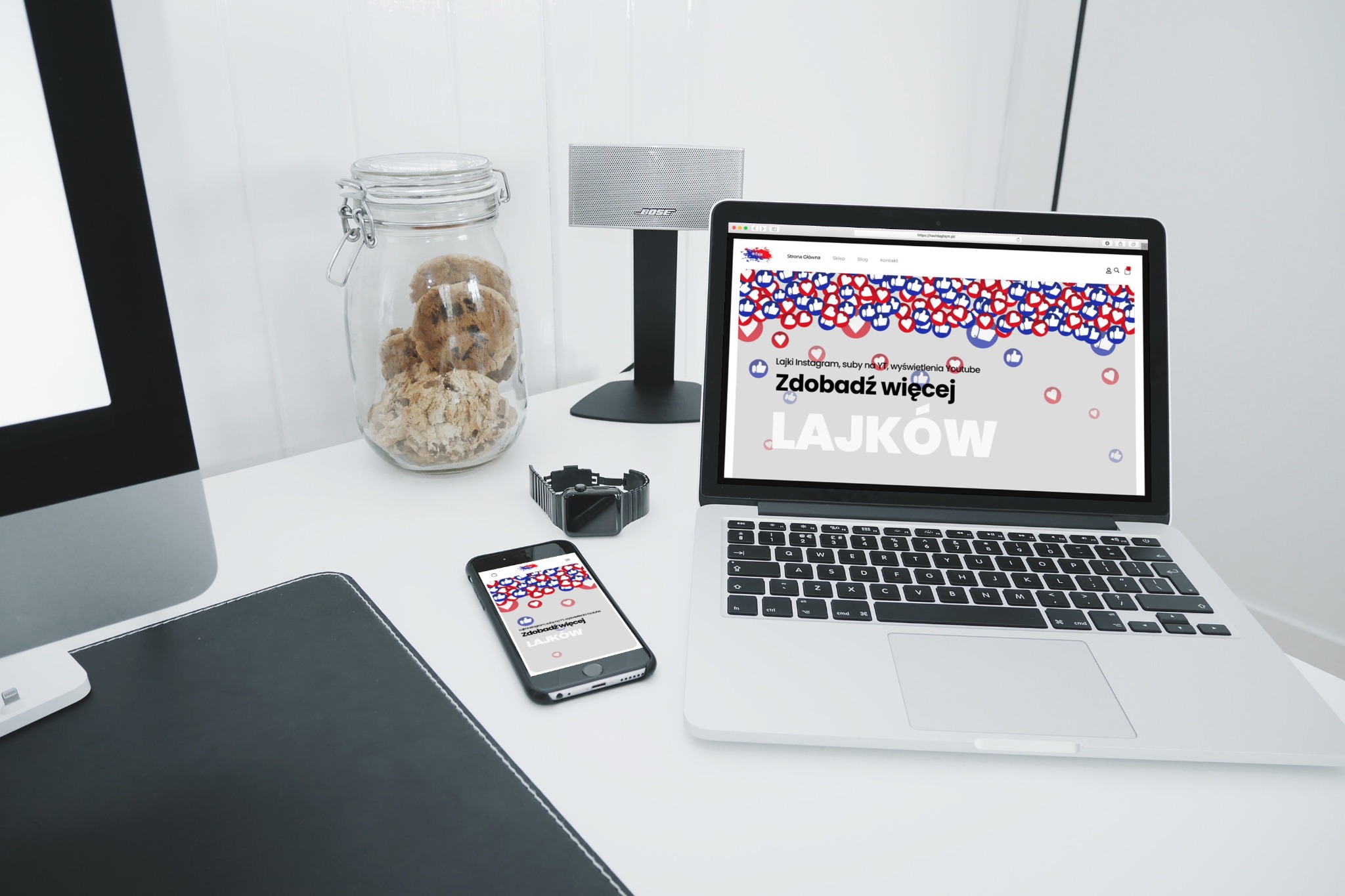 Laptop i smartfon na biurku prezentują stronę internetową z motywem polubień w kolorach czerwonym i niebieskim, obok słoik z ciasteczkami, głośnik Bose i smartwatch.