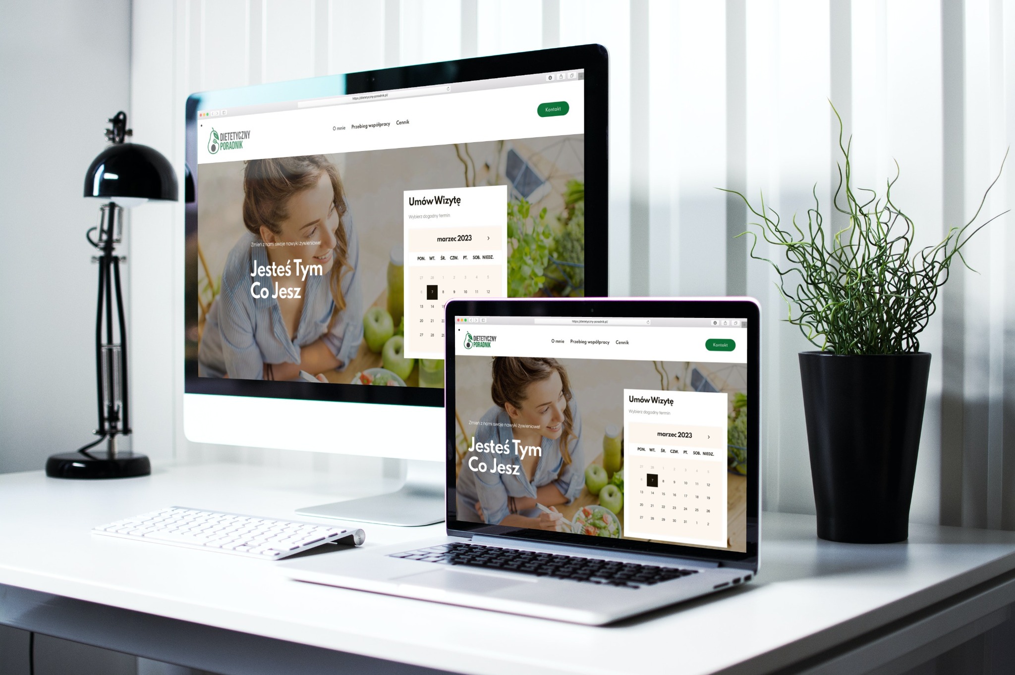 Ekran komputera stacjonarnego i laptopa prezentujące stronę internetową dietetyka z kalendarzem rezerwacji wizyt, obok klawiatura, lampka i roślina doniczkowa na białym biurku.