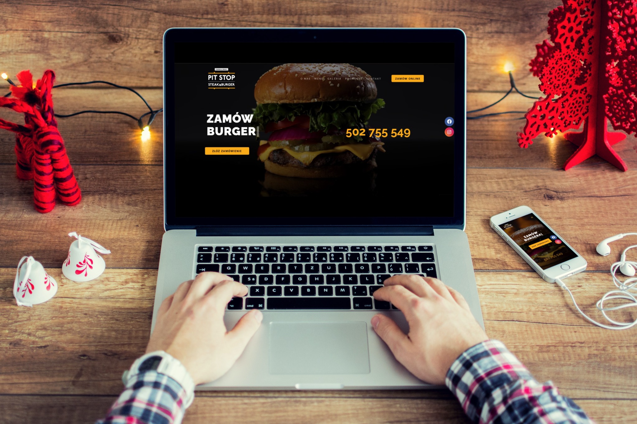 Osoba przegląda stronę internetową restauracji z burgerami na laptopie, obok leży smartfon z wyświetloną tą samą stroną, w tle drewniany stół z świątecznymi dekoracjami.