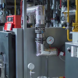 Szereg kotłów przemysłowych w kotłowni, z widocznymi rurami, zaworami, manometrami i panelami sterowania w kolorze czerwonym, otulonych folią izolacyjną.