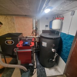 Dział EcoTerm firmy Strażak Sp. z o.o. - Nowoczesna instalacja grzewcza z czerwonym kotłem na suchy opał, czarnym zasobnikiem c.w.u. i rozdzielaczem Uponor, zamontowana w pomieszczeniu technicznym.