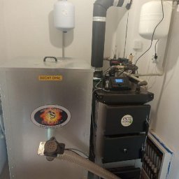 Dział EcoTerm firmy Strażak Sp. z o.o. - Zainstalowany piec na paliwo stałe 'eco DESIGN' z podajnikiem 'DRAGON' (SUCHY OPAŁ) w kotłowni. Widoczne izolowane rury, dwa naczynia wzbiorcze i panel sterowania.