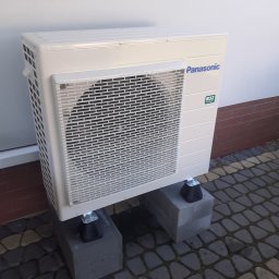 Dział EcoTerm firmy Strażak Sp. z o.o. - Zewnętrzna jednostka pompy ciepła Panasonic, zamontowana na dwóch betonowych bloczkach na kostce brukowej, przy białej ścianie budynku z czerwoną podmurówką.
