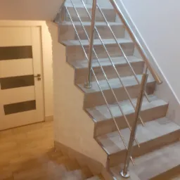 Schody wewnętrzne wykończone płytkami imitującymi drewno, z metalową balustradą i białymi ścianami. Widoczna także biała, nowoczesna drzwi z poziomymi przeszkleniami.