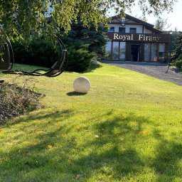 Royal Firany - Budynek firmy 'Royal Firany' z przeszkloną fasadą i brązowymi panelami, otoczony zadbanym trawnikiem z dekoracyjną kulą i wiszącymi fotelami.