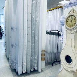 Royal Firany - Wystawa różnych wzorów firan i zasłon w sklepie, z dekoracyjnym, kremowym zegarem stojącym w tle po prawej stronie.