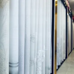 Ekspozycja pionowych, białych żaluzji materiałowych z różnymi wzorami haftów, wiszących w rzędzie w sklepie.