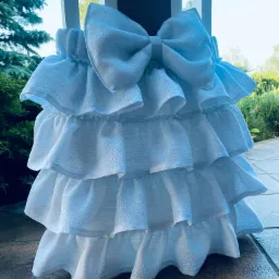 Biała roleta rzymska z trzema falbanami i dużą kokardą, widok z zewnątrz na tle zieleni.