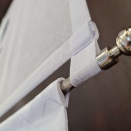 Royal Firany - Detale mocowania białej, półprzezroczystej rolety rzymskiej na chromowanym karniszu z ozdobną kulą na tle ciemnego mebla.