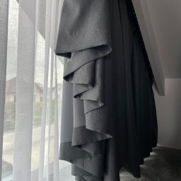 Royal Firany - Szara roleta rzymska z dekoracyjnymi zakładkami przy oknie z białą, zwiewną firaną w jasnym pokoju na poddaszu.