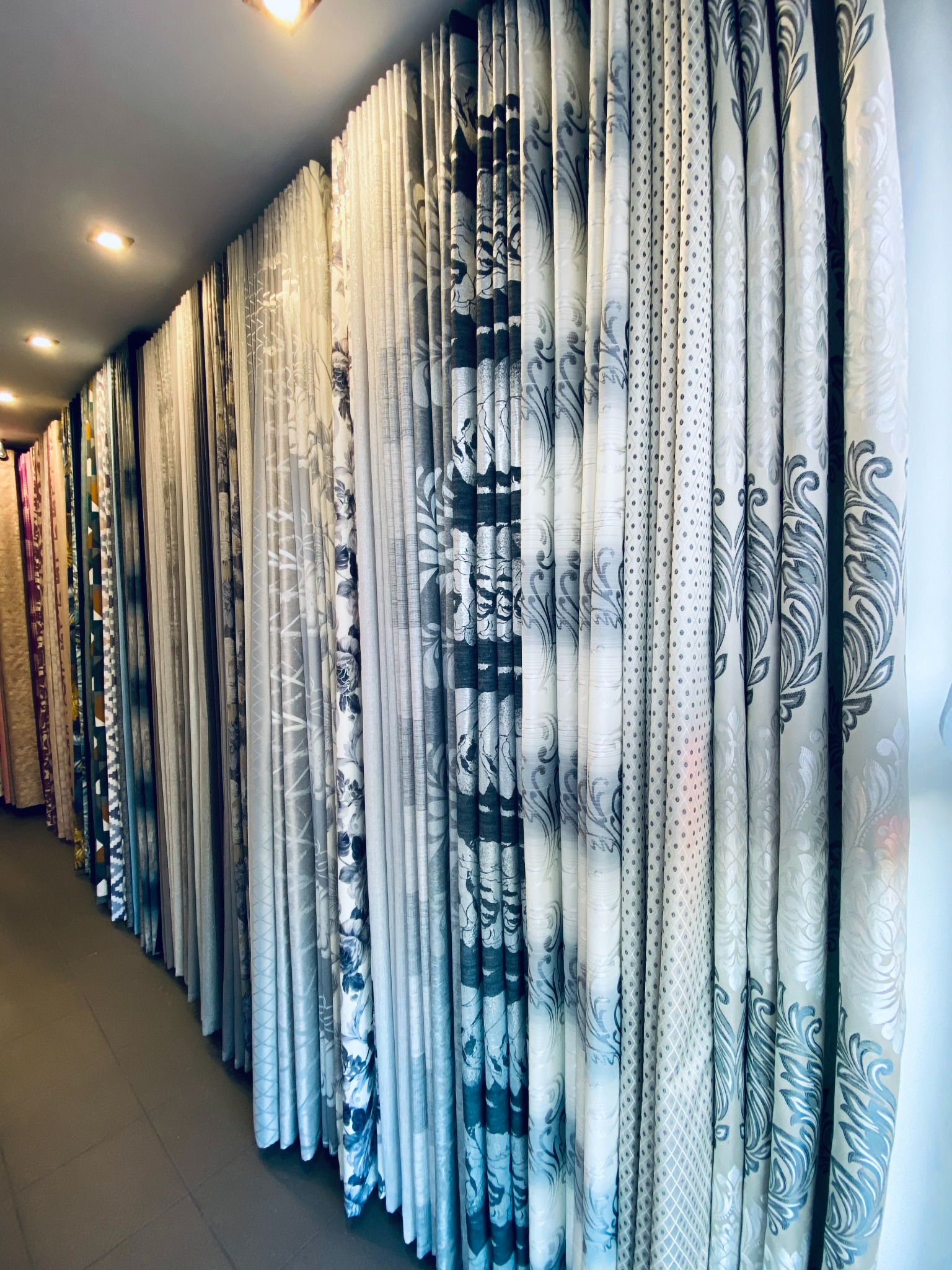 Różnorodne tkaniny zasłonowe z wzorami kwiatowymi, geometrycznymi i kropkami, prezentowane w rzędzie w sklepie z oświetleniem sufitowym.