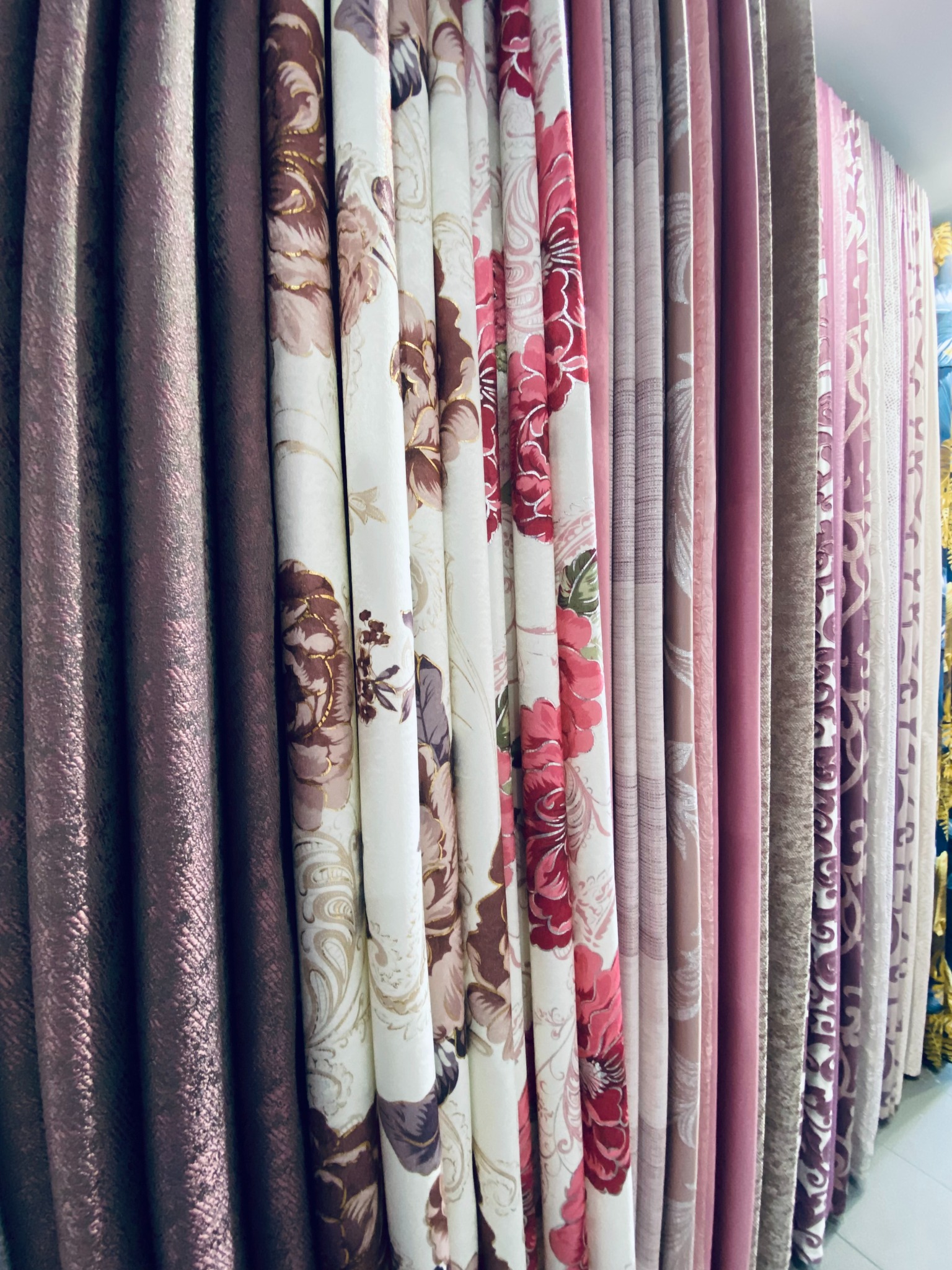 Różnorodność tkanin zasłonowych z motywami kwiatowymi i gładkimi, w odcieniach różu, brązu i beżu, prezentowanych w ekspozycji hurtowni.