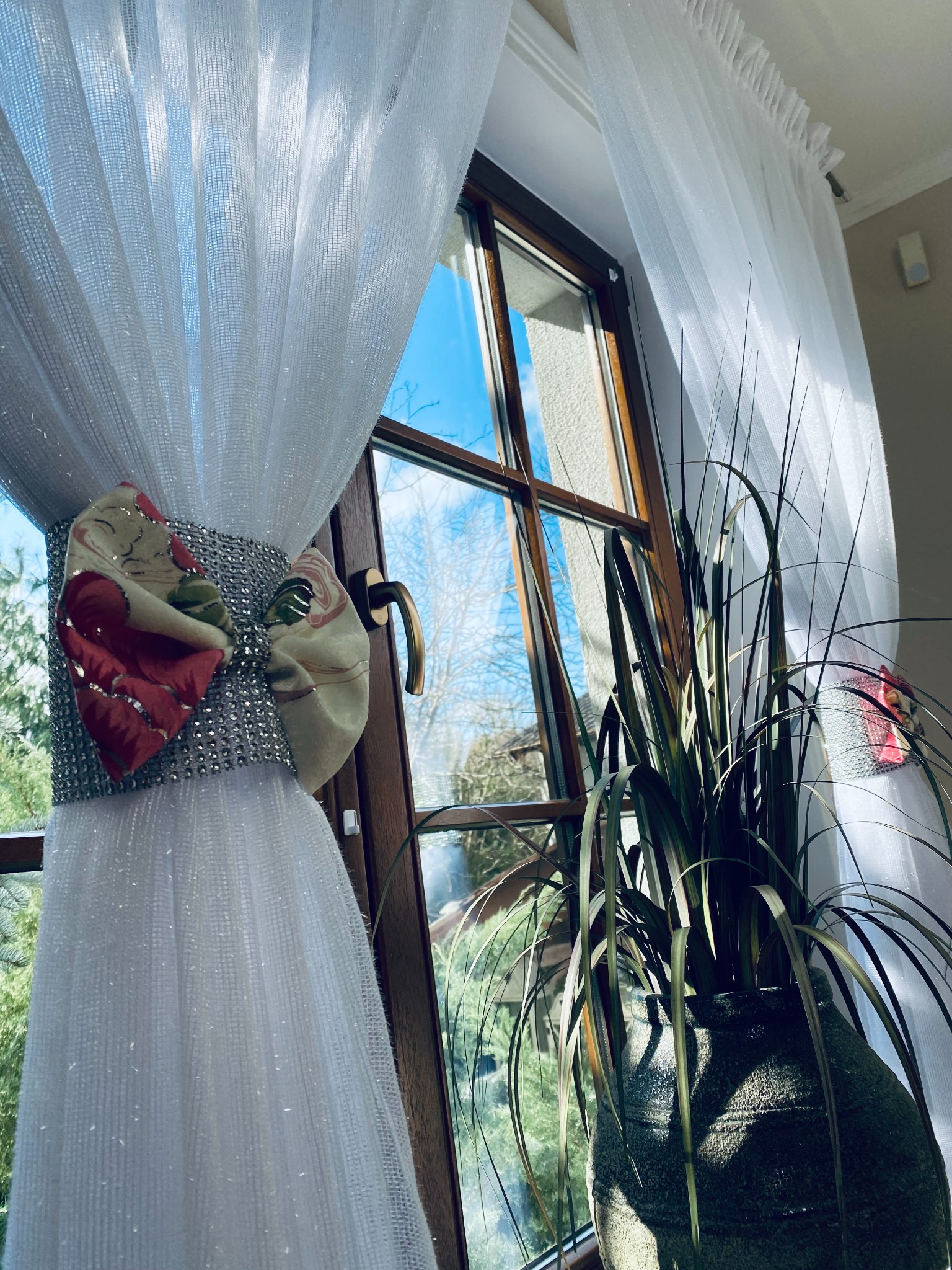 Przezroczysta, biała zasłona z kokardą z motywem kwiatowym, upięta na drewnianym oknie, obok doniczki z zieloną rośliną.