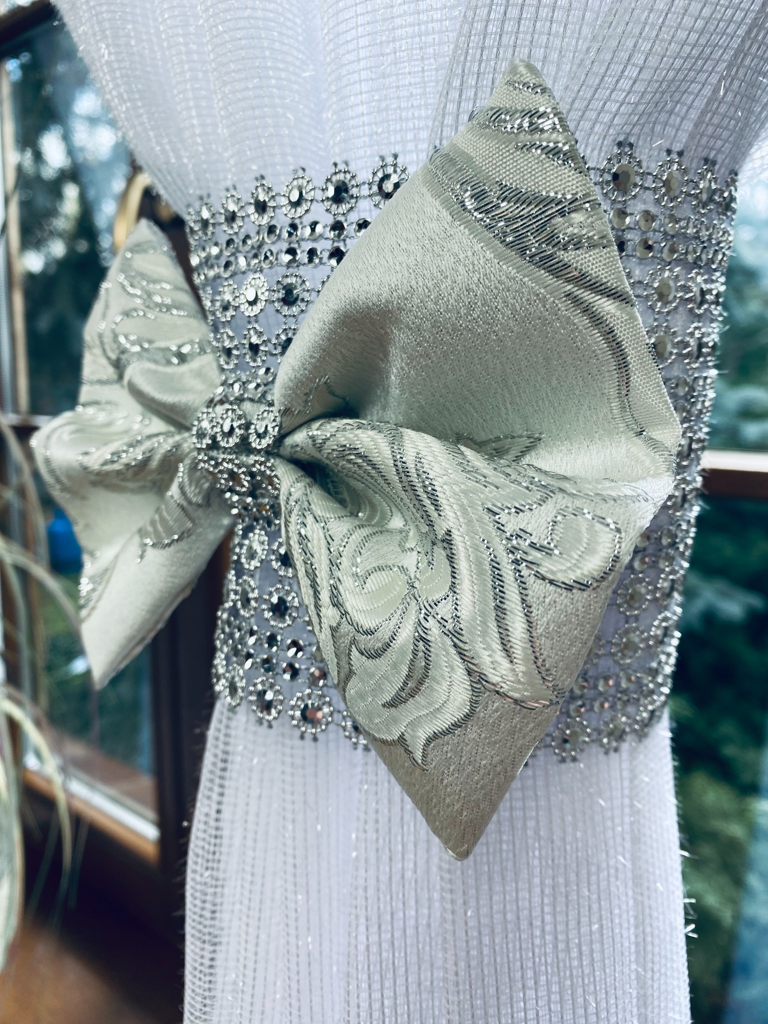 Biała, siateczkowa zasłona upięta srebrną taśmą z kryształkami i ozdobnym, srebrnym kokardą z wzorem w liście, widok z bliska na tle okna.