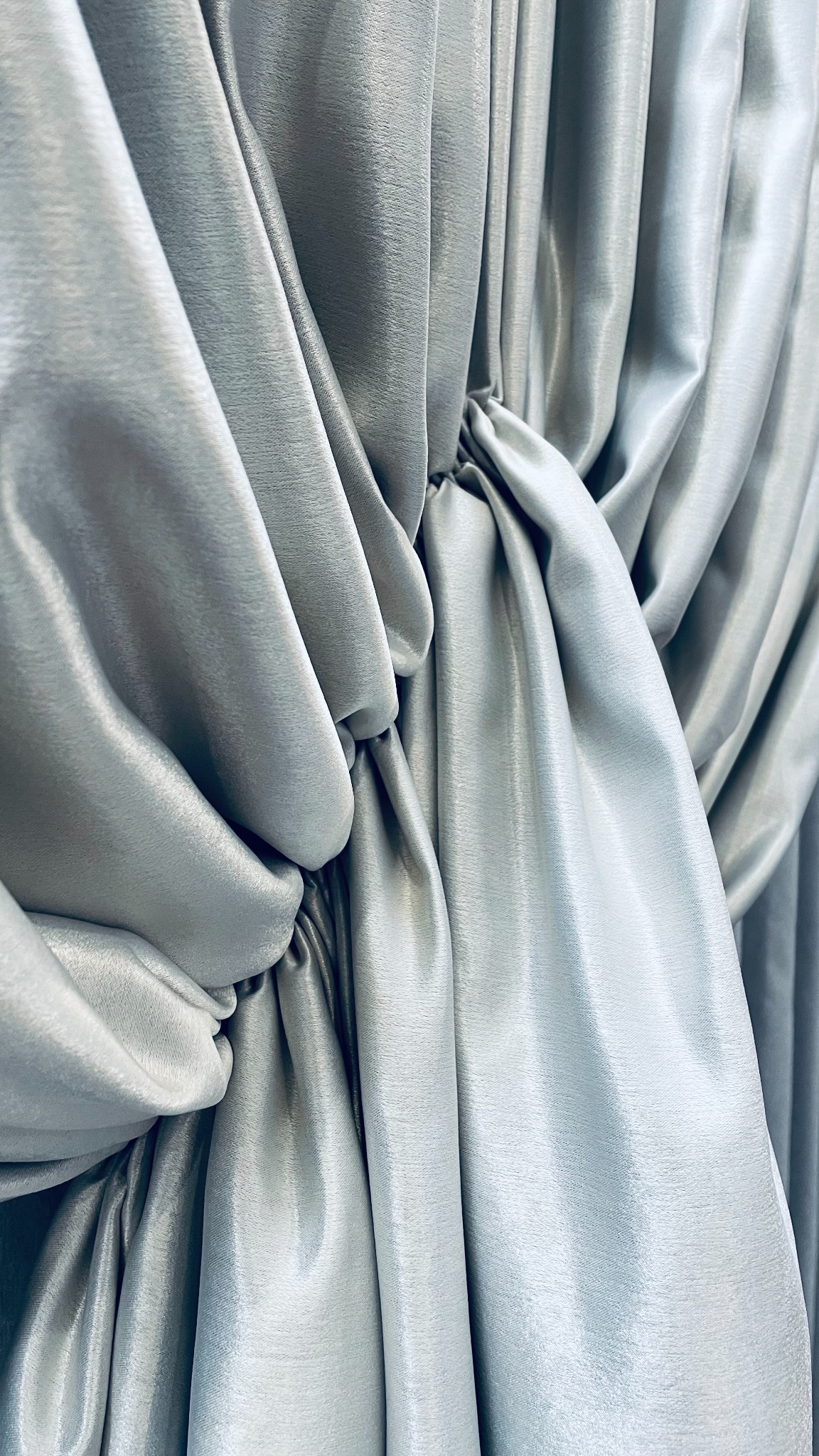 Szaro-srebrna, błyszcząca tkanina drapowana w dekoracyjny węzeł, tworząca miękkie fałdy i cienie.
