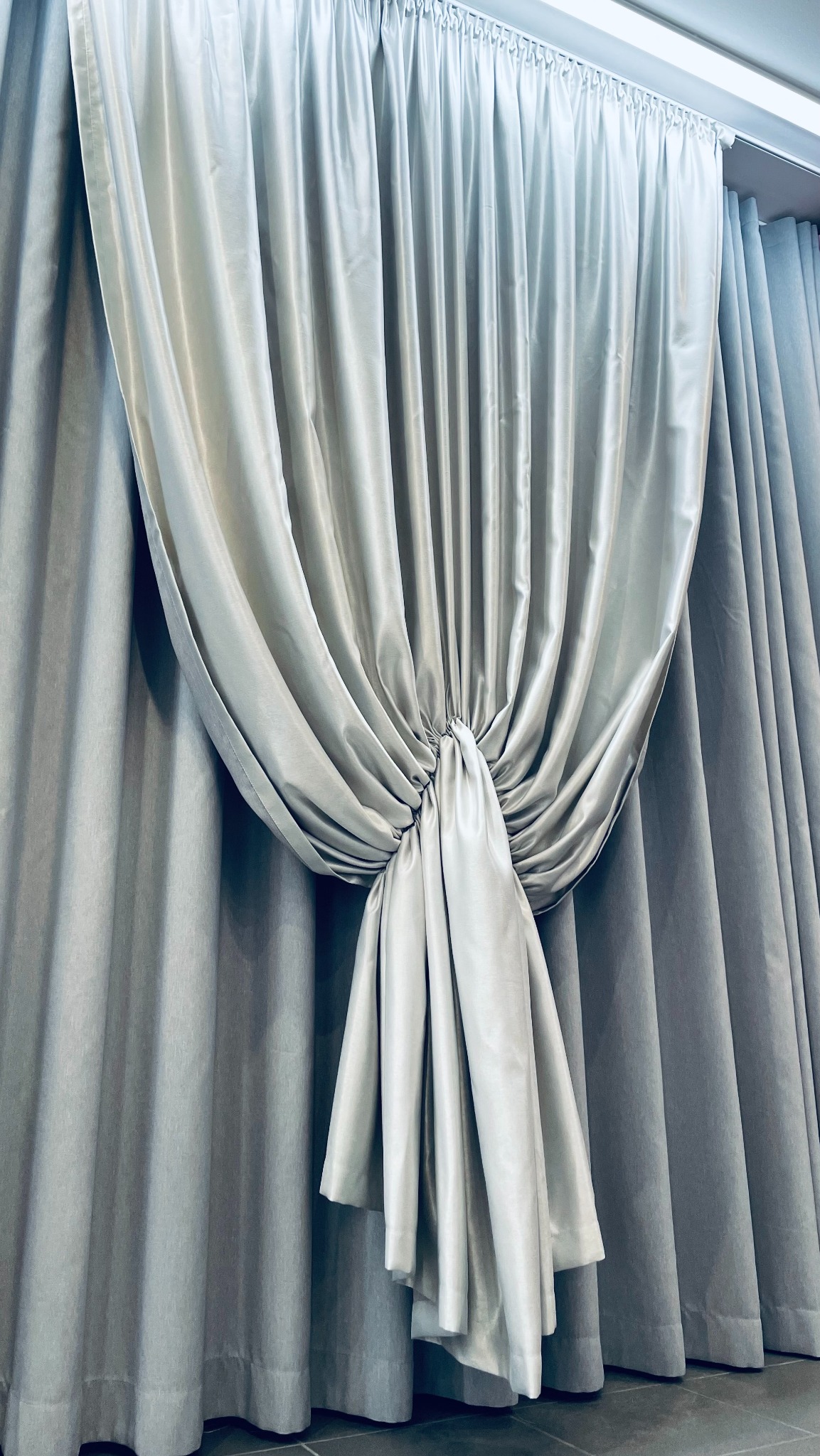 Elegancka, srebrzysta zasłona upięta w dekoracyjny węzeł, tworząca efektowny drapowany układ na tle gładkich, szarych zasłon.