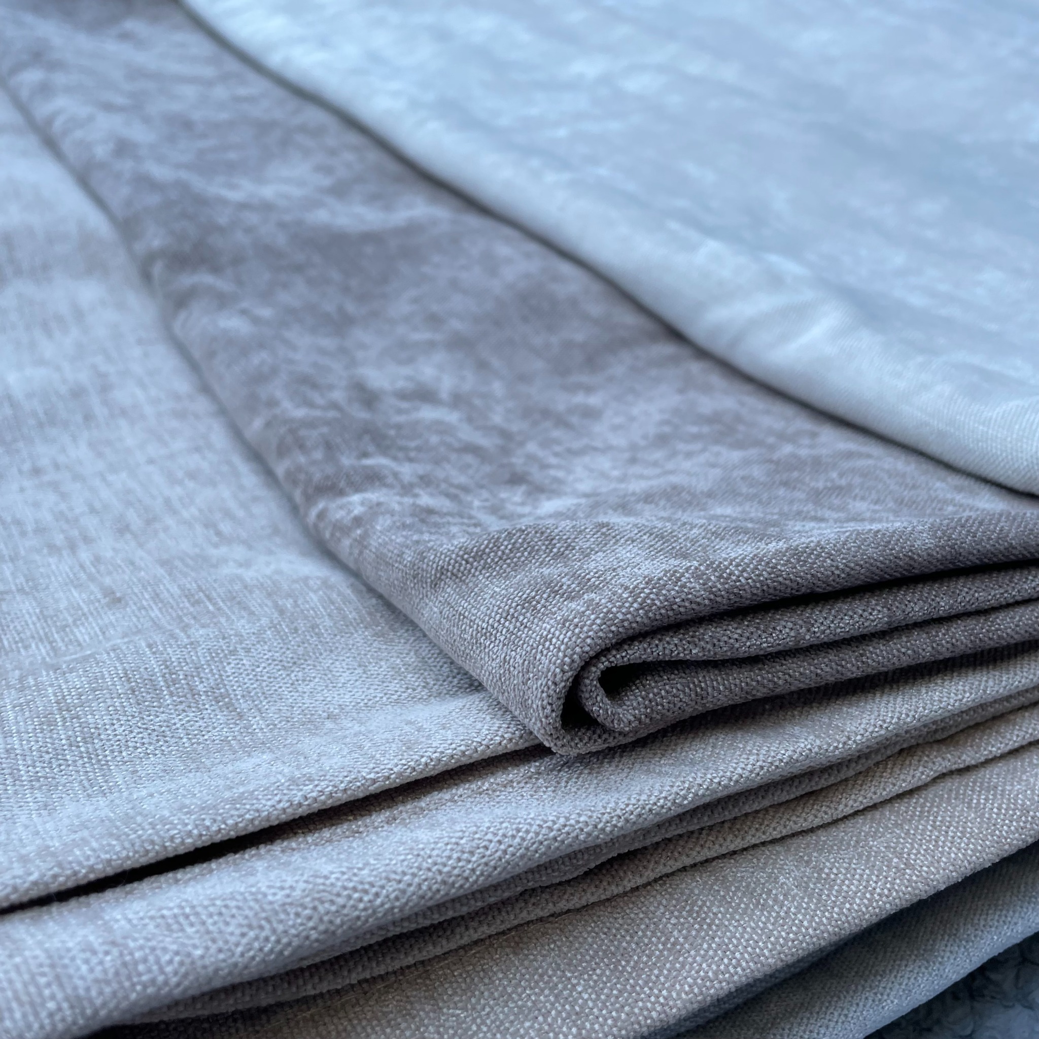 Stos trzech różnych tkanin w odcieniach szarości i beżu, z wyraźną fakturą i subtelnym połyskiem, ułożonych warstwami pod delikatnym kątem.