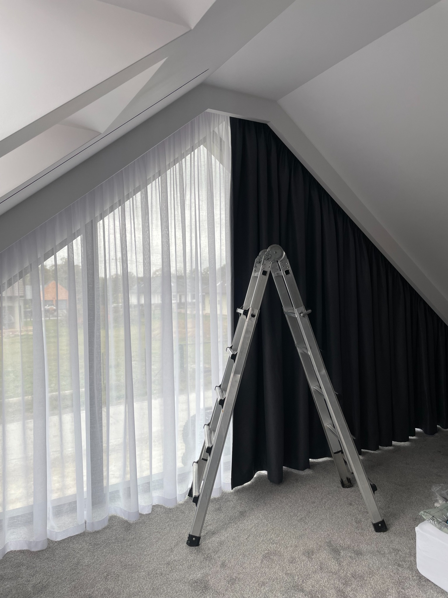 Ukończona instalacja białych, zwiewnych i czarnych, zaciemniających zasłon w pokoju na poddaszu ze skośnym sufitem. Aluminiowa drabina stoi na dywanie.
