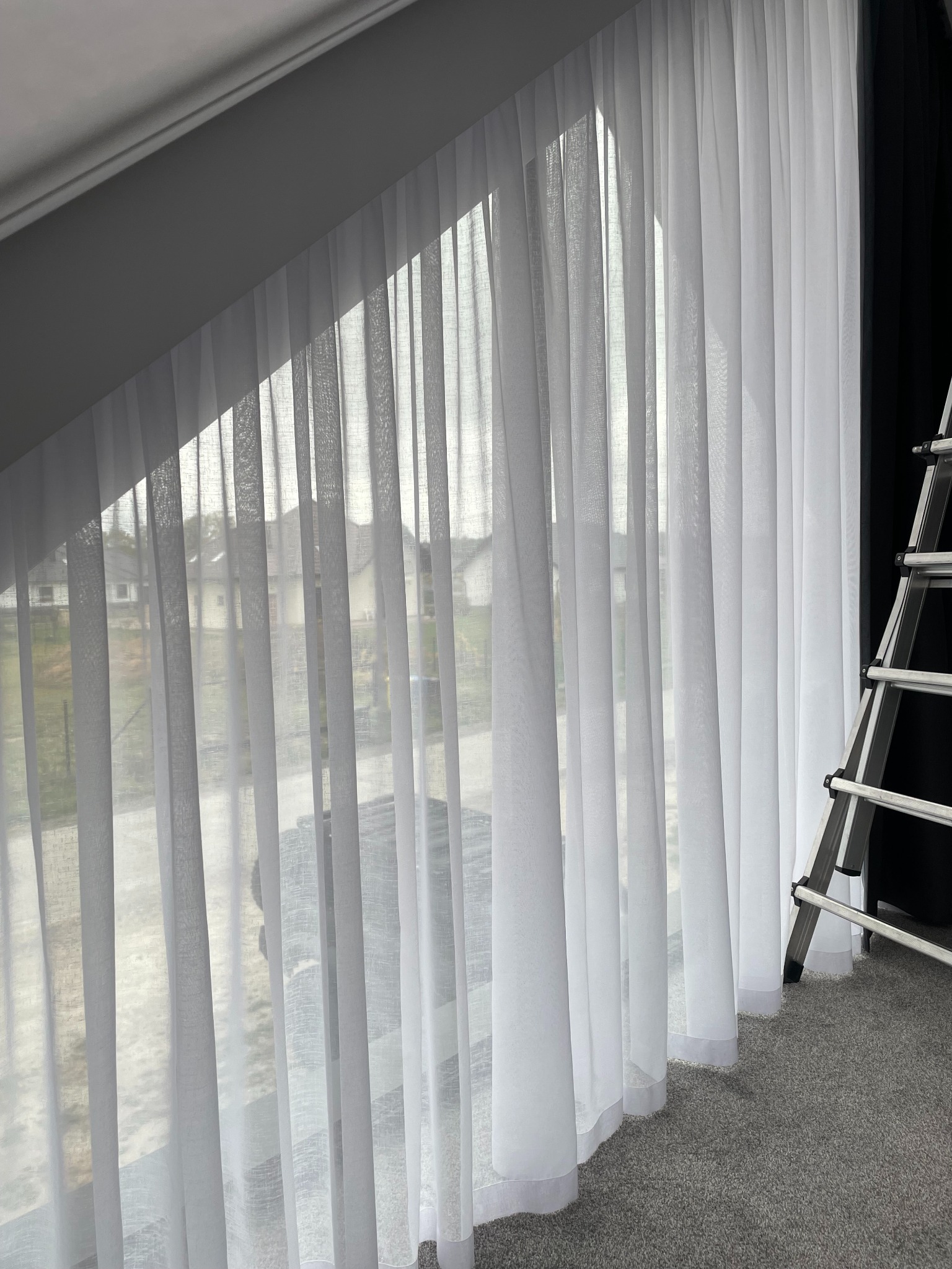 Delikatne, białe zasłony woalowe w oknie na poddaszu, z widokiem na domy. Aluminiowa drabina oparta o ścianę. Subtelna elegancja w aranżacji okiennej.