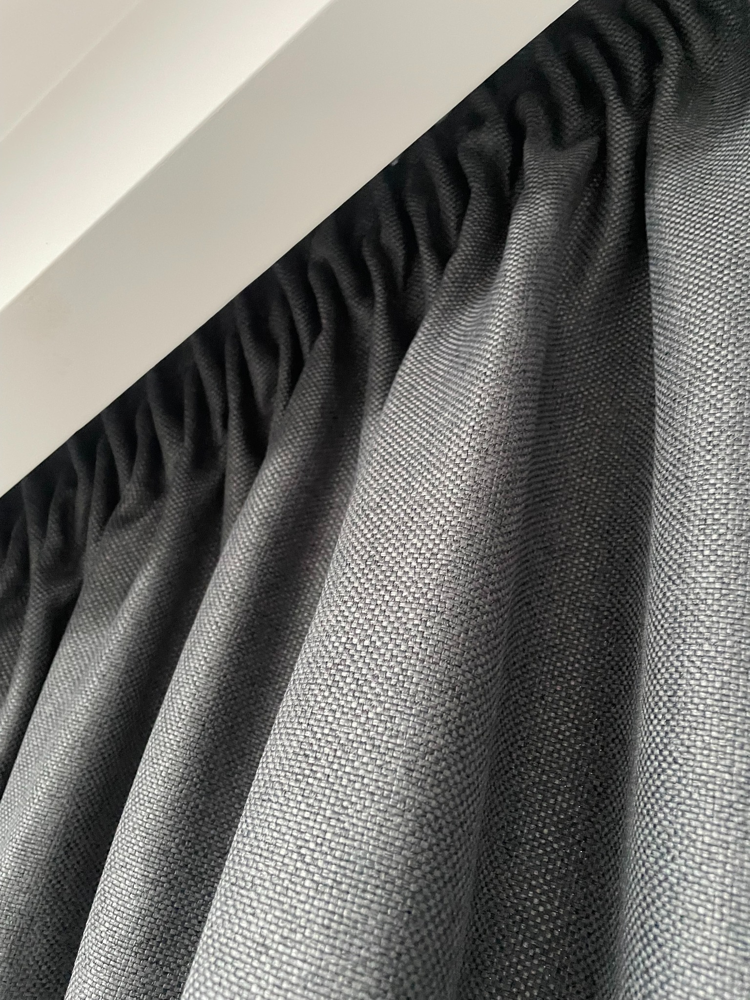 Szara, teksturowana zasłona z marszczeniem u góry, zawieszona na białym karniszu. Widoczny detal splotu materiału i subtelne fałdy.