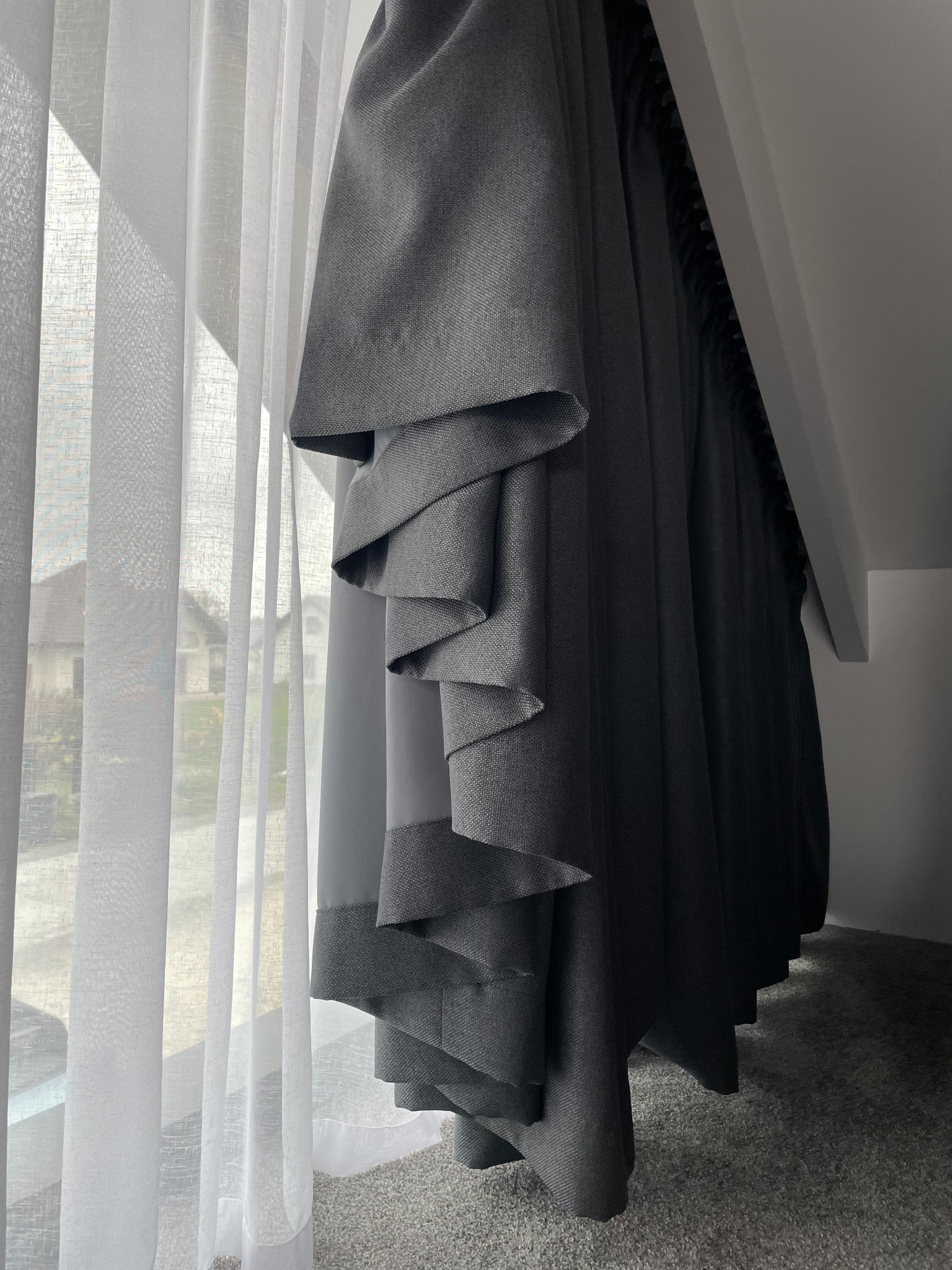 Szara roleta rzymska z dekoracyjnymi zakładkami przy oknie z białą, zwiewną firaną w jasnym pokoju na poddaszu.