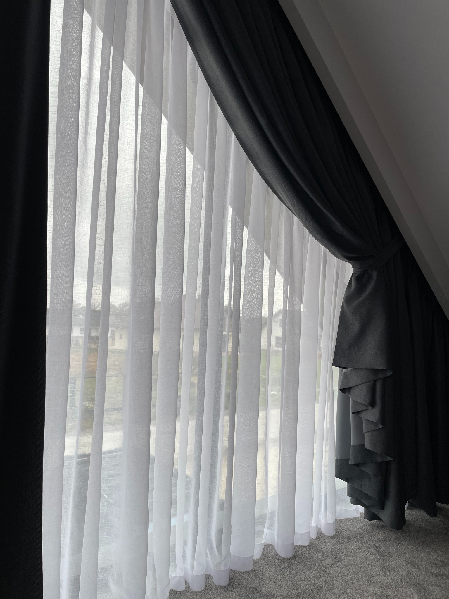 Okno na poddaszu z białymi, delikatnymi firanami i grubymi, grafitowymi zasłonami upiętymi z boku. Widok na zewnątrz przez firany.