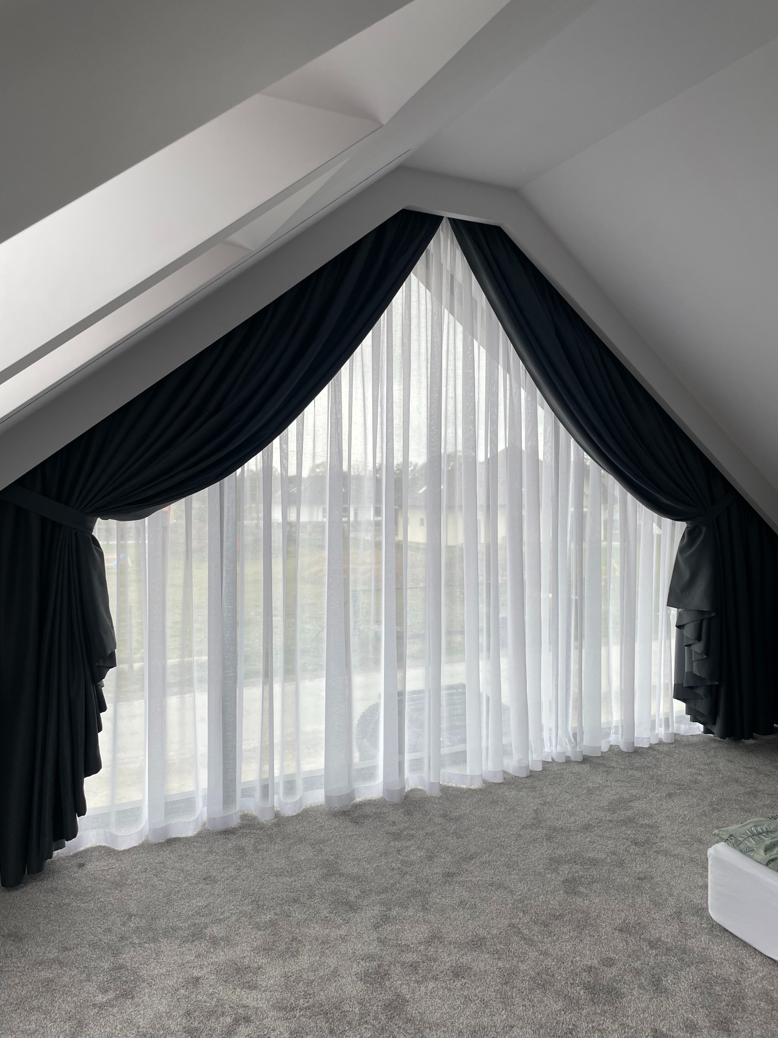 Zasłony zaciemniające i firany w oknie na poddaszu. Ciemne zasłony upięte po bokach okna, przez środek prześwitująca firana. Szara podłoga z wykładziny dywanowej.
