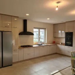 Jasna, nowoczesna kuchnia z beżowymi, lakierowanymi frontami, drewnianymi akcentami i czarnym okapem, widok na dwudrzwiową lodówkę Samsung i stół z zielonym blatem.