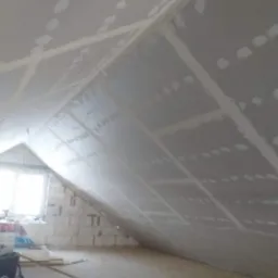 Poddasze w trakcie wykańczania płytami gipsowo-kartonowymi, widoczne łączenia płyt zaszpachlowane, okno w szczycie ściany z pustaków.