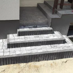 Schody wejściowe do domu w trakcie budowy, wyłożone szarą kostką brukową, widoczne narzędzia na górnym stopniu i piasek wokół.