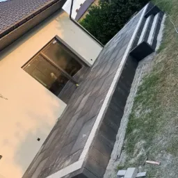 Nowoczesny taras z szarych płyt betonowych z białymi obrzeżami w trakcie budowy, widoczne niezabudowane płyty i narzędzia na trawie obok budynku.