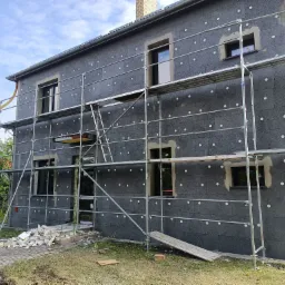 Dom w trakcie ocieplania, ściany pokryte szarym styropianem z białymi kołkami, widoczne rusztowania i resztki materiałów budowlanych na ziemi.