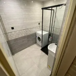 Wykończona łazienka z pralką, szklaną kabiną prysznicową z czarnymi ramami i umywalką na szafce, płytki ścienne w odcieniach szarości z geometrycznym wzorem, widok zza otwartych drzwi.