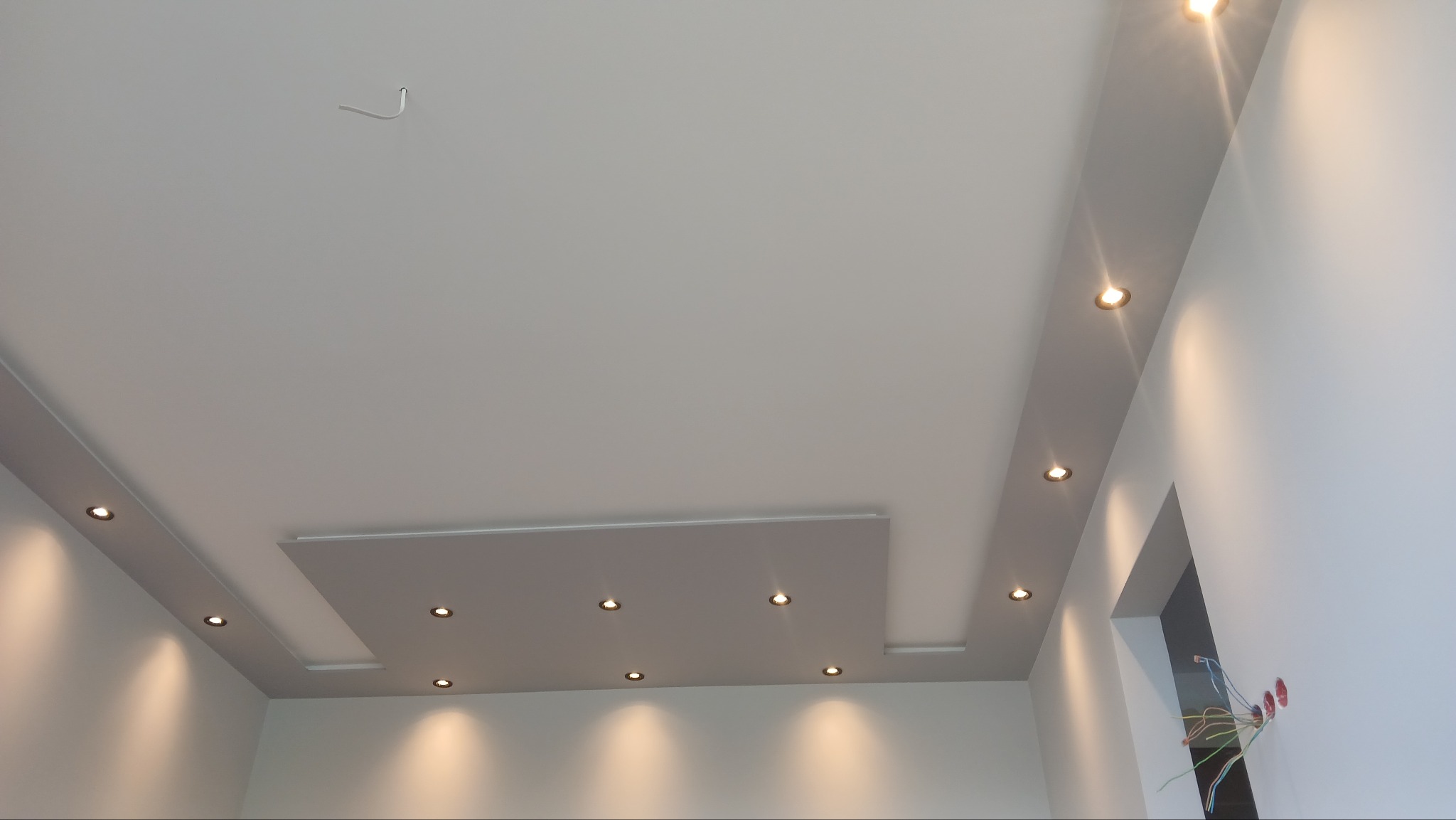 Nowoczesny sufit podwieszany z oświetleniem punktowym, wykończony w odcieniach bieli i szarości. Widoczne przewody elektryczne w ścianie, sugerujące etap prac wykończeniowych.