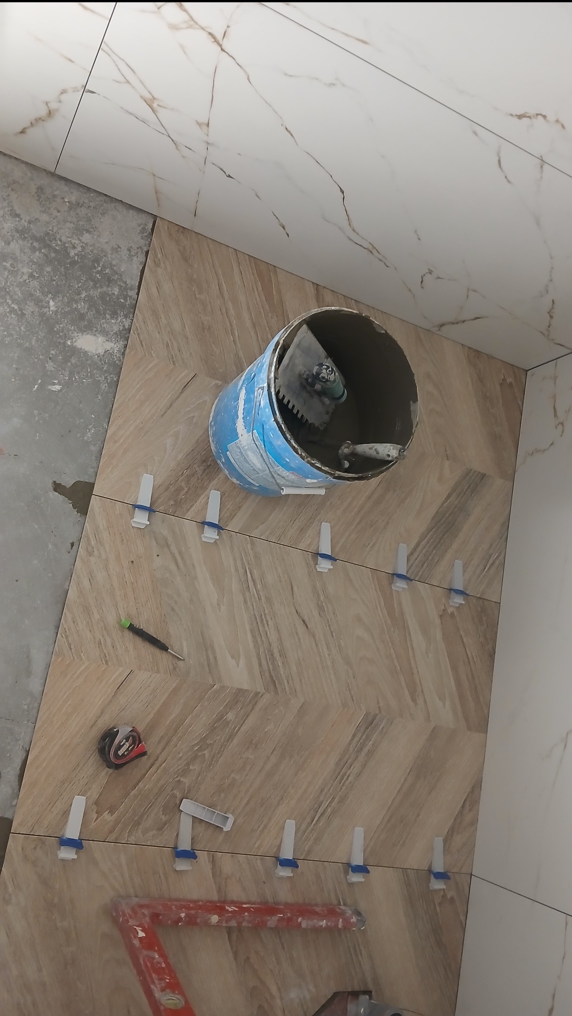 Układanie płytek imitujących drewno w jodełkę, z użyciem poziomicy, krzyżyków dystansowych i wiadra z zaprawą. Częściowo wykończona ściana z białych płytek.