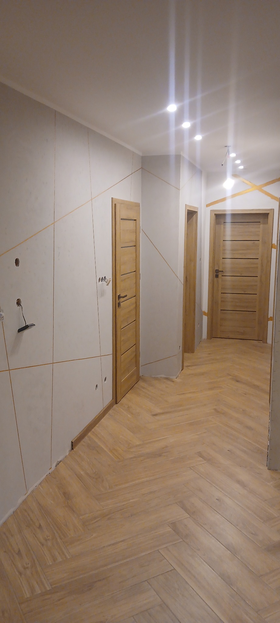 Korytarz w trakcie wykańczania: jasne ściany z dekoracyjnymi liniami, drewniane drzwi i podłoga ułożona w jodełkę. Widoczne niedokończone prace przy ścianach.