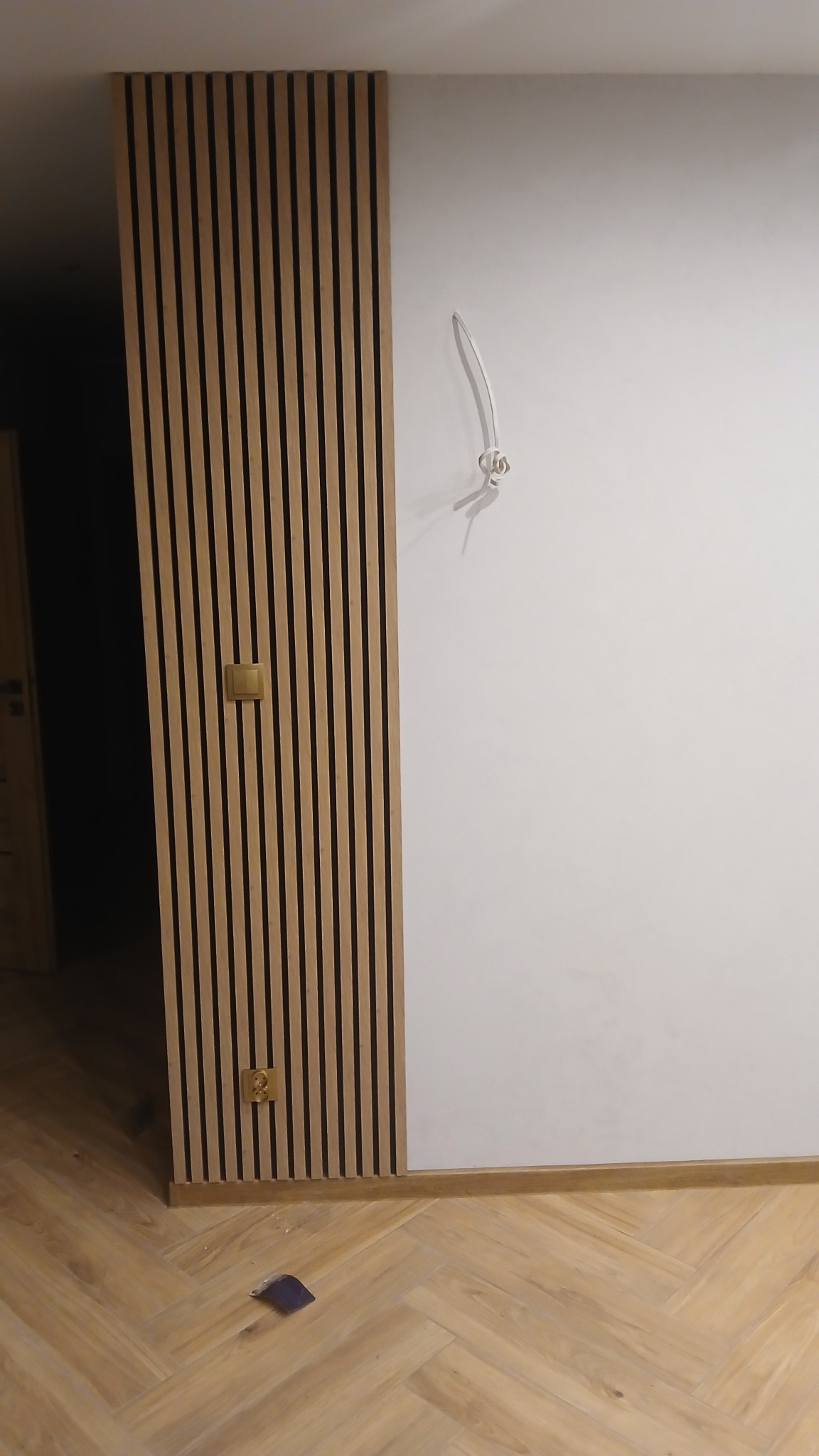 Panel dekoracyjny z drewnianych listewek na ścianie, z widocznym gniazdkiem i wystającym kablem. Parkiet ułożony w jodełkę na podłodze.