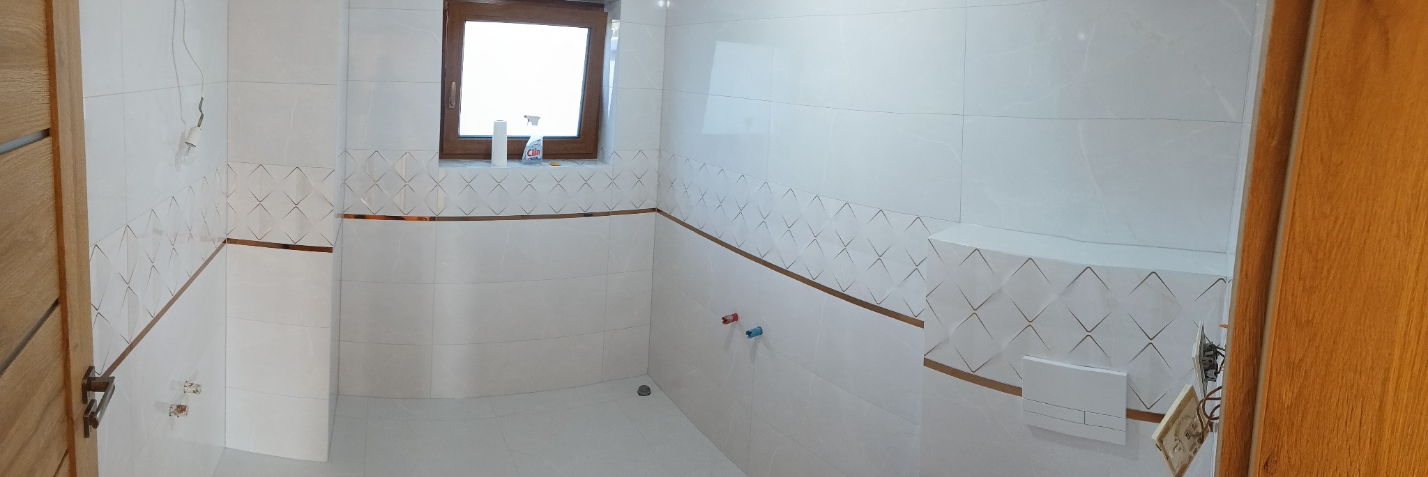 Wykończona łazienka z białymi płytkami, dekoracyjnym pasem z motywem rombów i złotymi akcentami. Widoczne okno, armatura i fragment drewnianych drzwi.