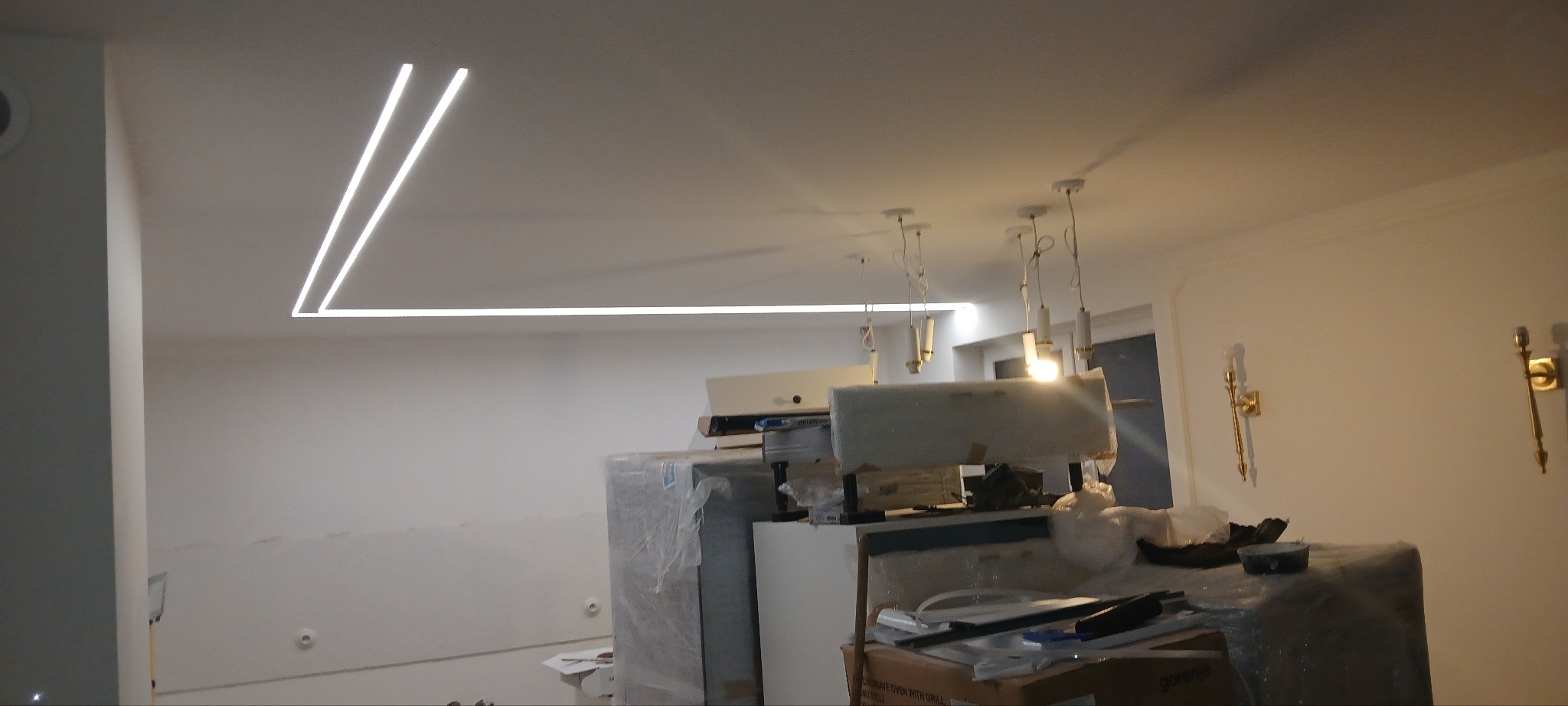 Nowoczesne oświetlenie sufitowe LED w kształcie litery L, wiszące lampy i kinkiety w remontowanym wnętrzu. Widoczne materiały budowlane.