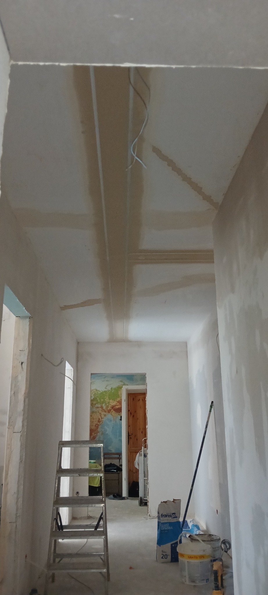 Korytarz w trakcie prac wykończeniowych: widoczne łączenia płyt na suficie, drabina, worki z materiałami. W tle pomieszczenie z mapą świata na ścianie.