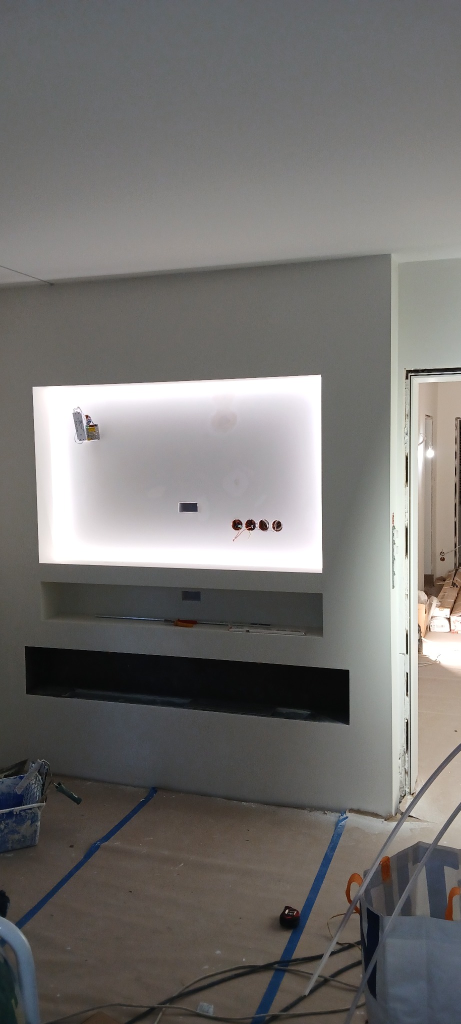 Nowoczesna ściana z podświetlaną wnęką i miejscem na TV, widoczne gniazdka elektryczne, w tle otwarte drzwi, podłoga zabezpieczona folią malarską.