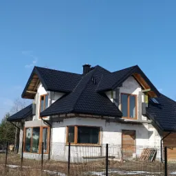 Dom jednorodzinny w trakcie budowy, z czarnym dachem z dachówki modułowej i oknami z ramami w kolorze drewna, widziany z zewnątrz, z elementami rusztowania i częściowo ośnieżonym terenem.