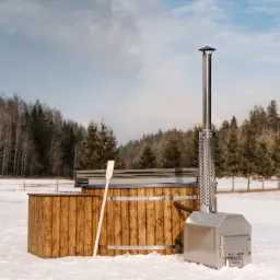 Drewniana sauna beczka z piecem na drewno i kominem na tle zimowego krajobrazu z lasem i śniegiem.