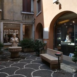 Foto Pablo Paweł Osiński - Dziedziniec w Wenecji z fontanną w kształcie kobiety, na tle witryn sklepowych i kamiennej ławki.