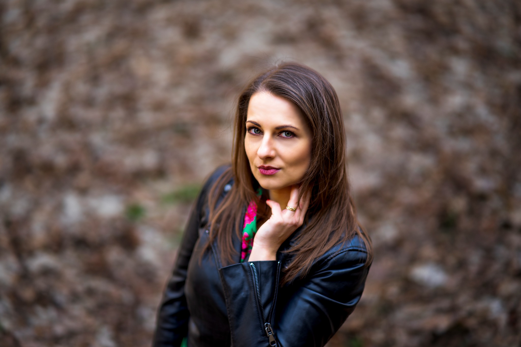 Portret kobiety w czarnej skórzanej kurtce, z zieloną apaszką w kwiaty, pozującej na tle rozmytego, brązowego tła.