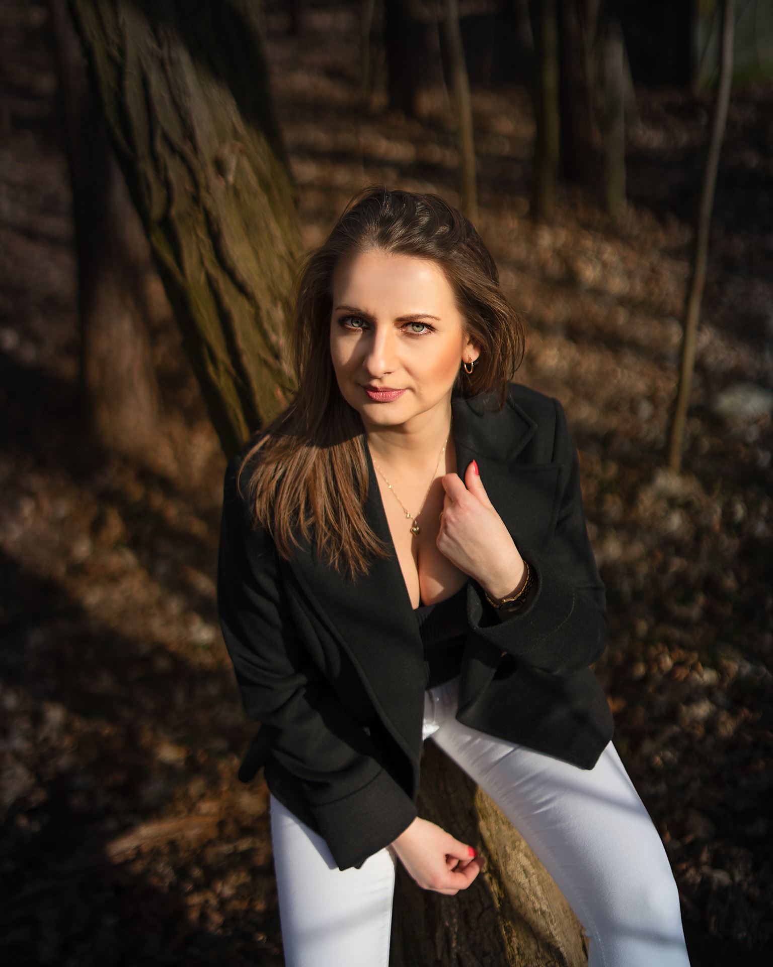 Portret kobiety w czarnym płaszczu i białych spodniach, opierającej się o drzewo w lesie, z delikatnym uśmiechem i złotą biżuterią.