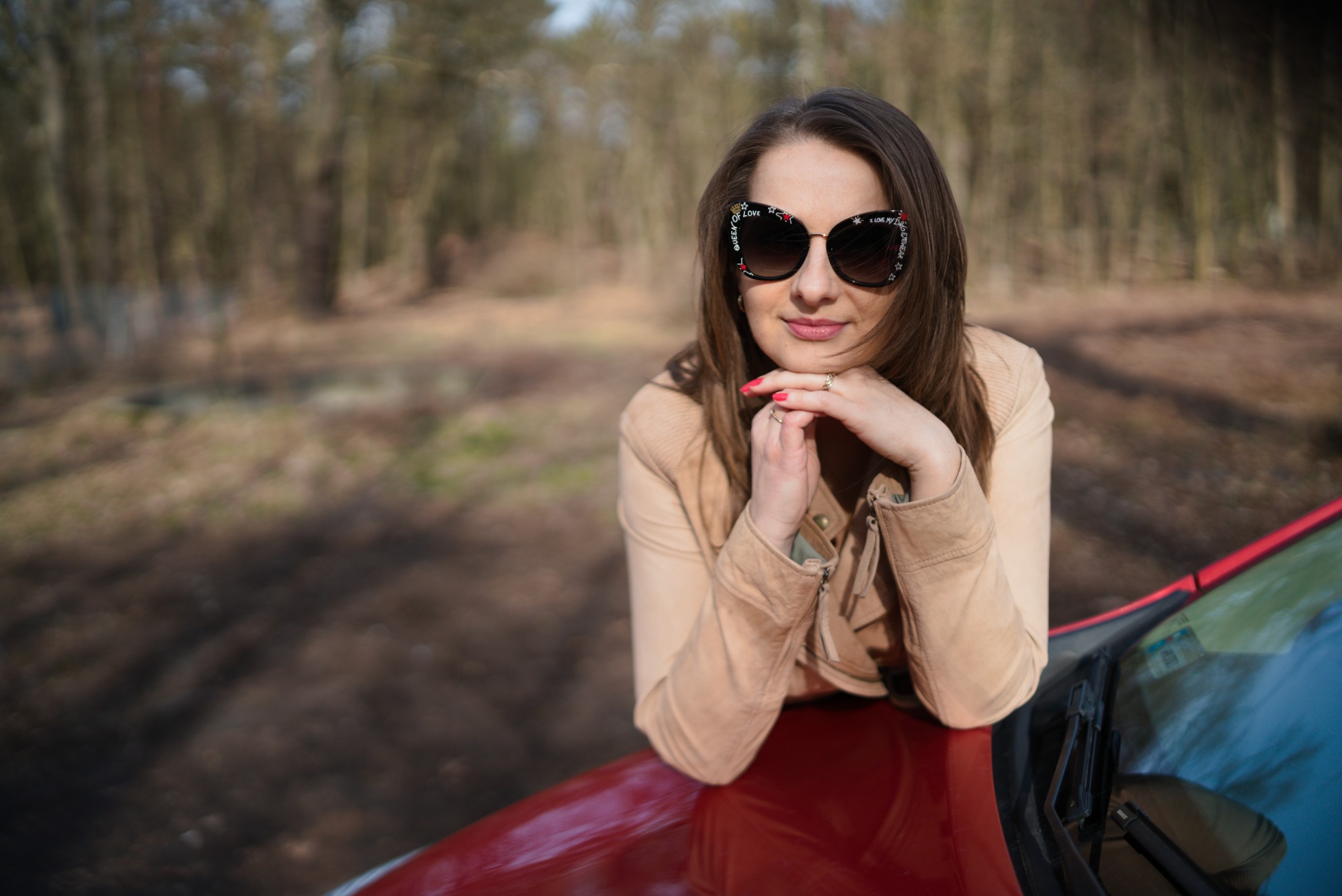 Portret kobiety w okularach przeciwsłonecznych z ozdobami, opierającej się o czerwony samochód na tle leśnym.