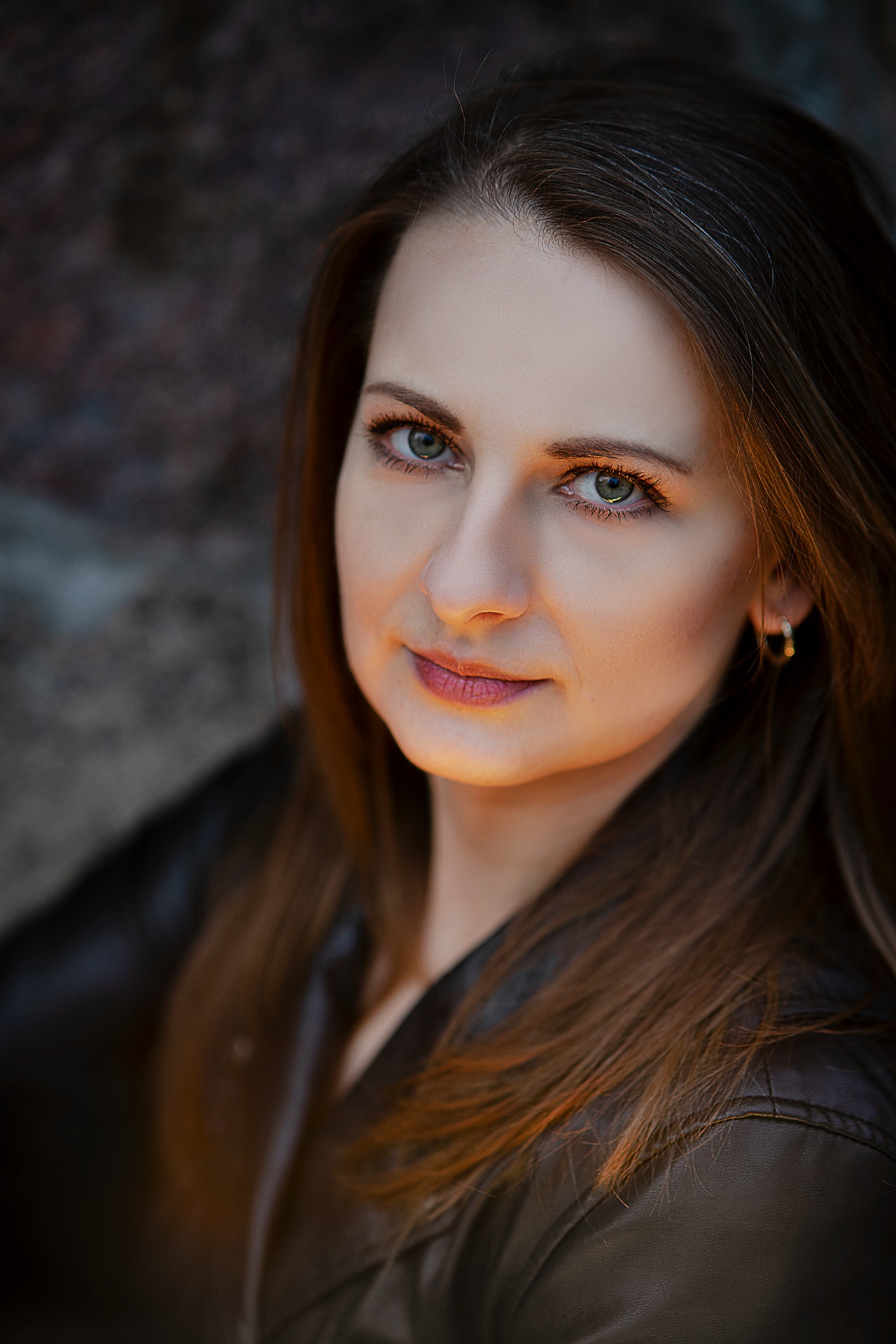 Portret kobiety z długimi brązowymi włosami, ubranej w skórzaną kurtkę, z subtelnym uśmiechem i zielonymi oczami, sfotografowanej z dołu na tle szarej ściany.