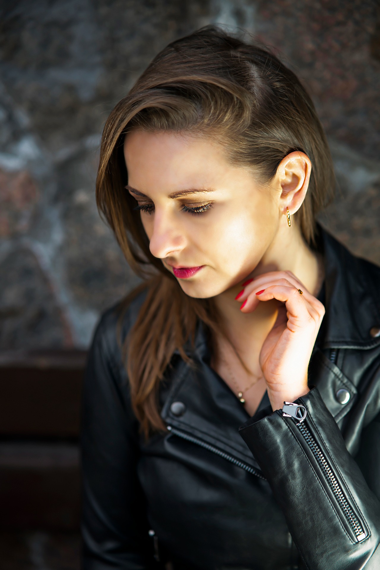 Portret kobiety w czarnej skórzanej kurtce, z delikatnym makijażem i złotą biżuterią, spoglądającej w dół, na tle kamiennej ściany.